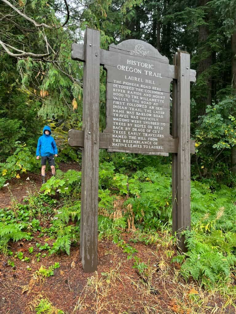 Our son stands on a hill behind the sign for Laurel Hill. The sign reads: "HISTORIC OREGON TRAIL
LAUREL HILL
THE PIONEER ROAD HERE DE TOURED THE COLUMBIA RIVER RAPIDS AND MOUNT HOOD TO THE WILLAMETTE VALLEY. THE ROAD AT FIRST FOLLOWED AN OLD INDIAN TRAIL. THE LATER NAME WAS BARLOW ROAD.
TRAVEL WAS DIFFICULT.
WAGONS WERE SNUBBED TO TREES BY ROPES OR HELD BACK BY DRAGS OF CUT TREES. EARLY TRAVELERS NAMED THE HILL FROM THE RESEMBLANCE OF NATIVE LEAVES TO LAUREL."
