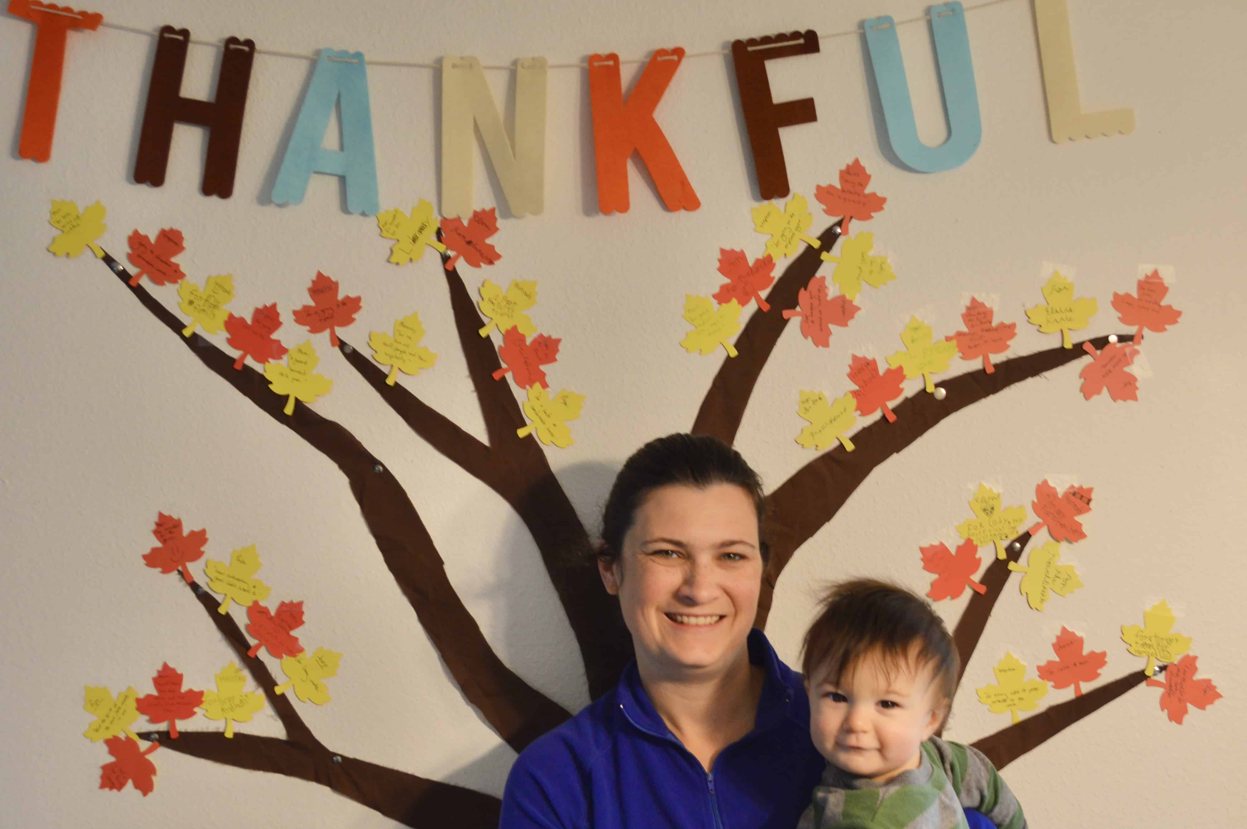 woman and baby in front of tree with paper leaves on wall with word "thankful" 