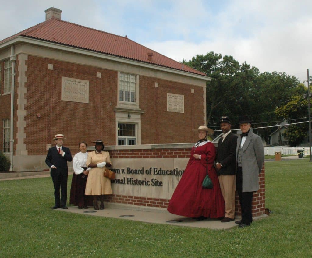 Six people dressed as living history characters stand beside the entrance sign for Brown v. Board of Education National Historic Site.