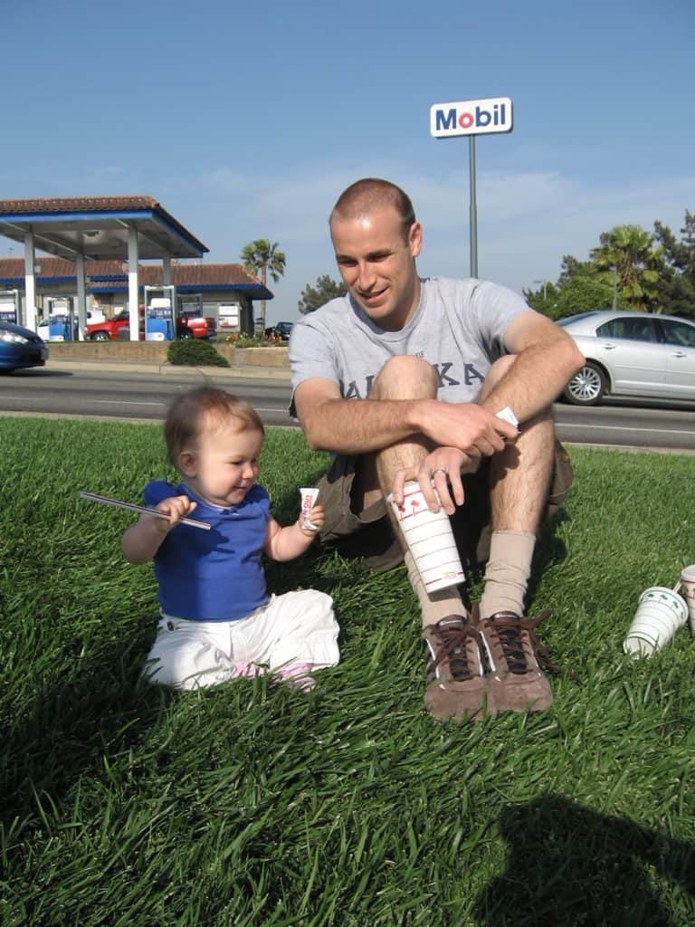 baby on grass with straw next to dad. On a road trip, sometimes the only green grass is near the road. Check out our road trip packing list printable for more tips!