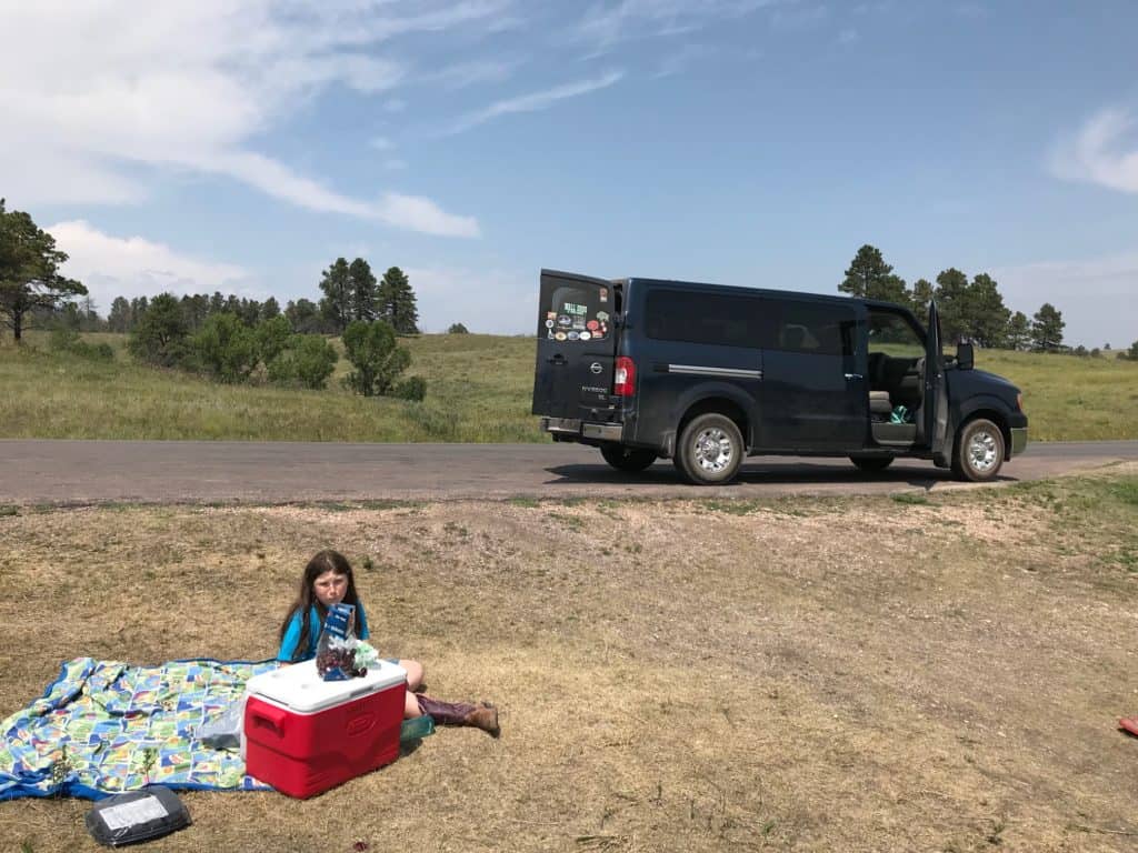 Girl on picnic blanket off side of road with van in background. Picnics are one of the best ideas for a road trip lunch.