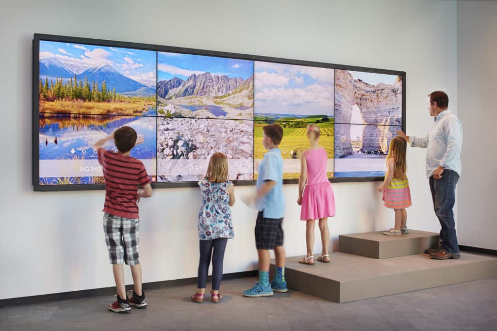 Children and a man play on touch screens to watch dinosaurs in various settings.