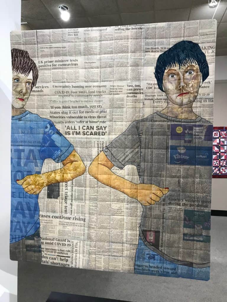 Quilt showing two people elbow greeting with headlines of the COVID-19 pandemic as background. Quilt from the National Quilt Museum