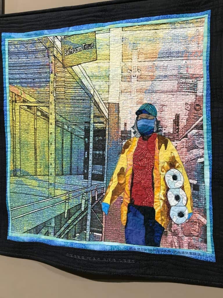 Quilt showing image of a woman picking up a package of toilet paper with empty shelves nearby. 