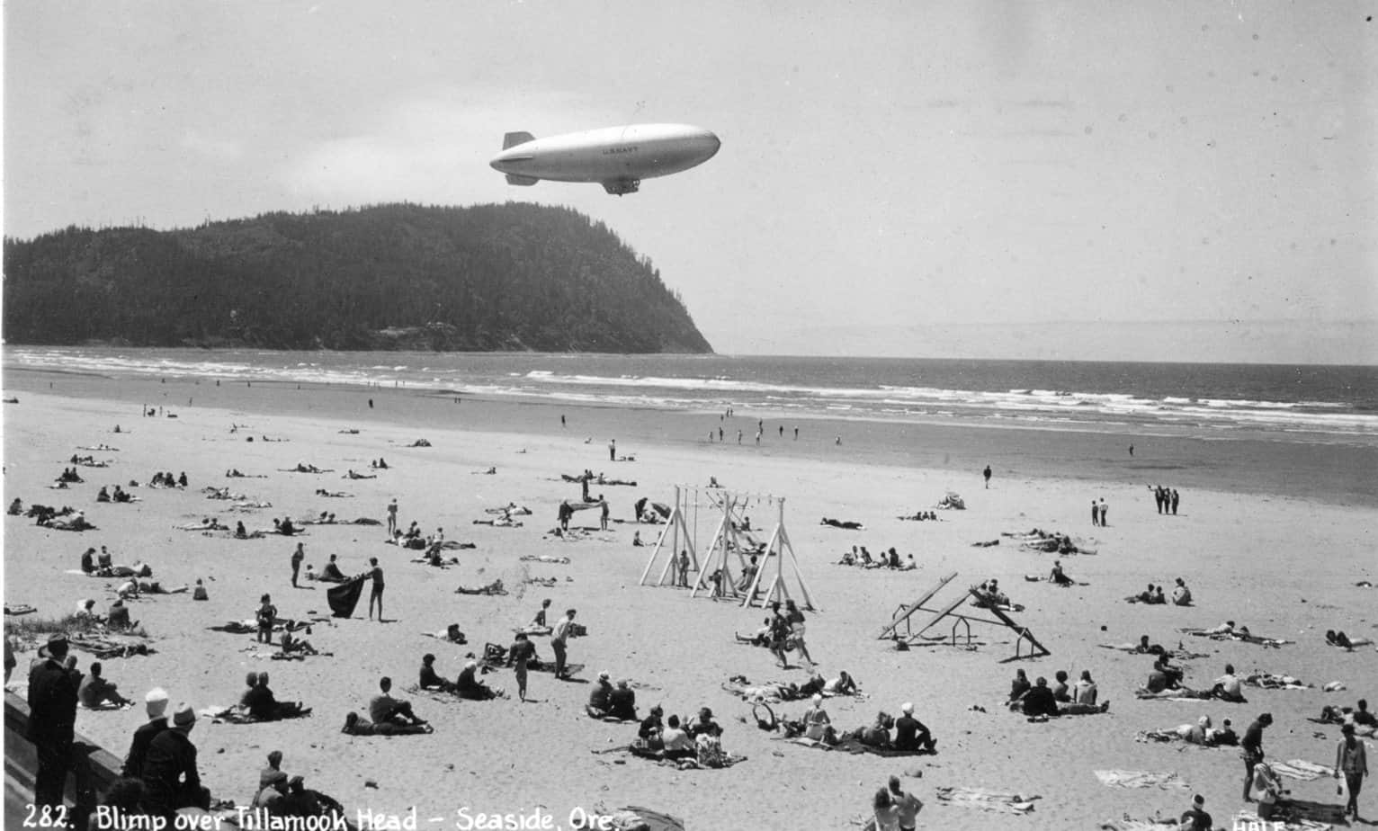 The black and white photo shows beachgoers at Seaside Beach. Several of them are looking up a K-Class blimp ascending over the beach.