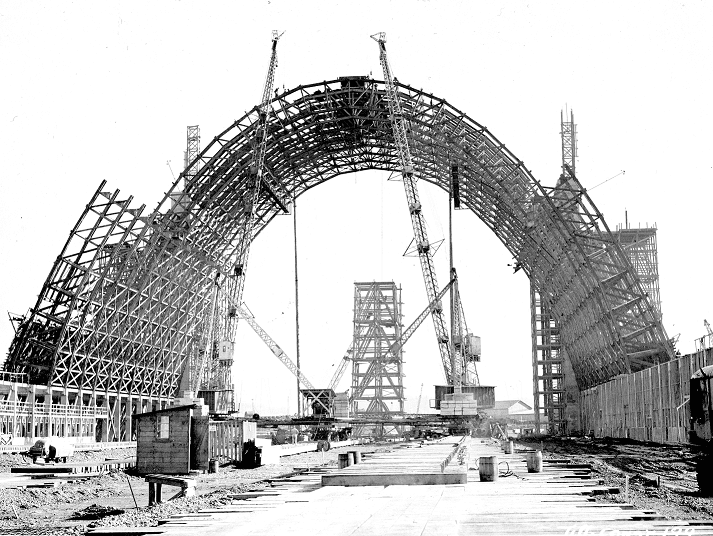 The black and white photo shows the construction of massive arch that makes up the roof and walls of Hangar B.
