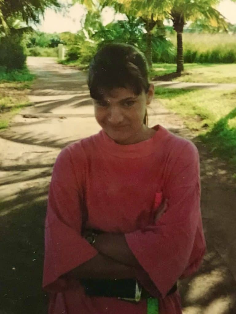 Teen girl with folded arms across chest and coconut palms in background. Anxiety can start early. This post contains several self-care ideas for anxiety.