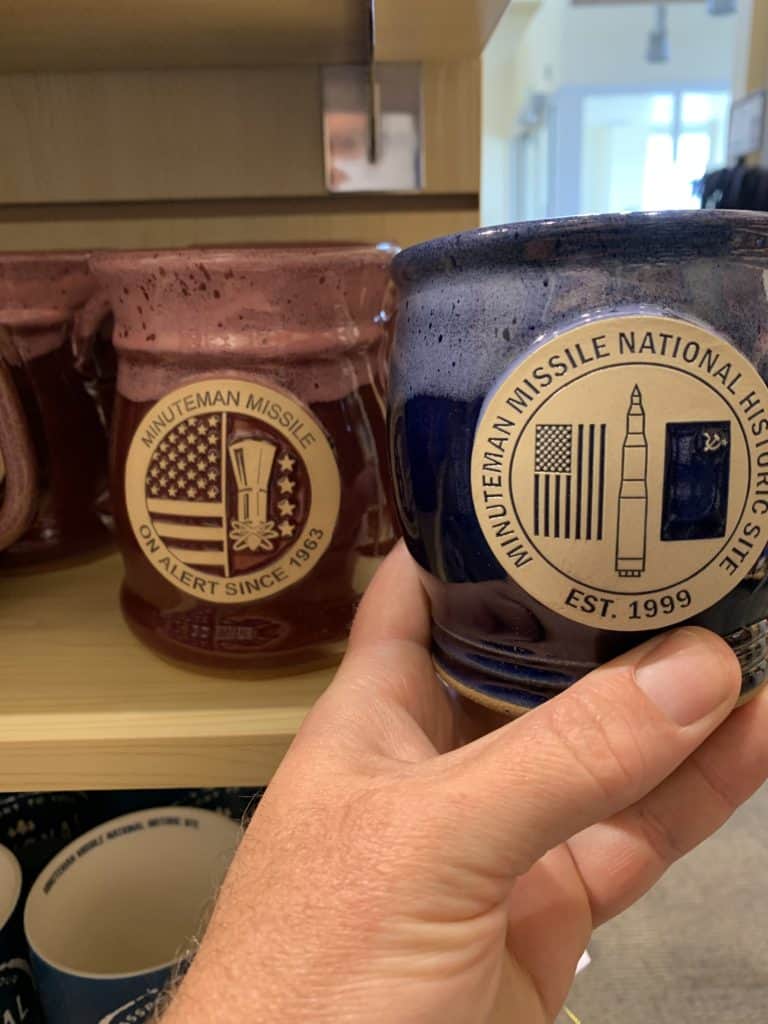 The Visitor Center shelf displays a variety of Daneen mugs created for Minuteman Missile NHS. My hand holds beautiful blue mug.