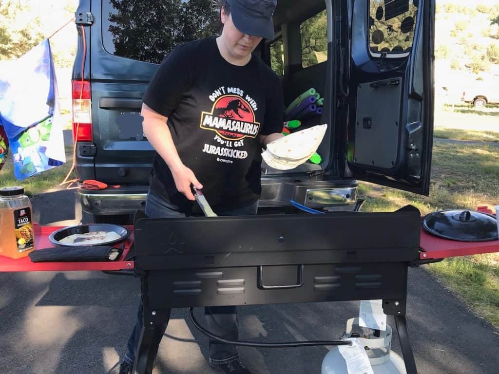 Mom cooking at camp stove in front of van. Keep cooking simple on your camping trip wherever possible.