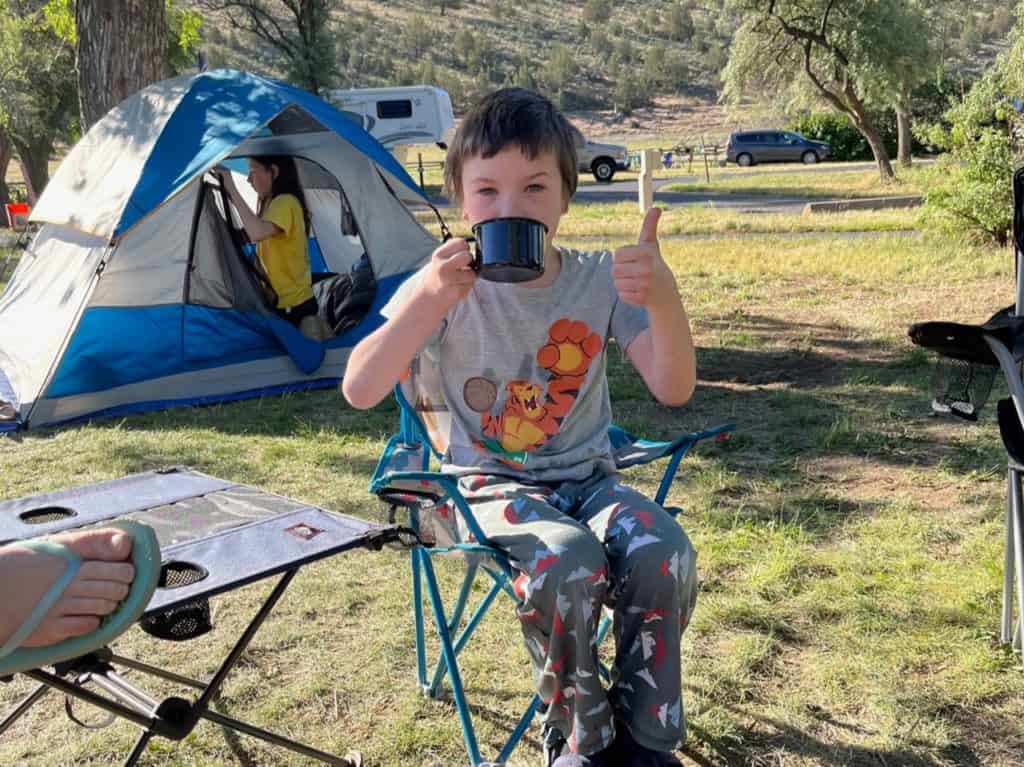 Boy with mug in camp chair giving the thumbs-up. We got some great camping ideas for families on our first camping trip!