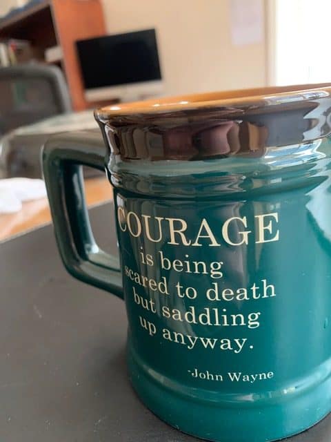 A tall green coffee mug bear the John Wayne quote: "Courage is being scared to death but saddling up anyway." Western mugs are some of the best cowboy gifts for men.