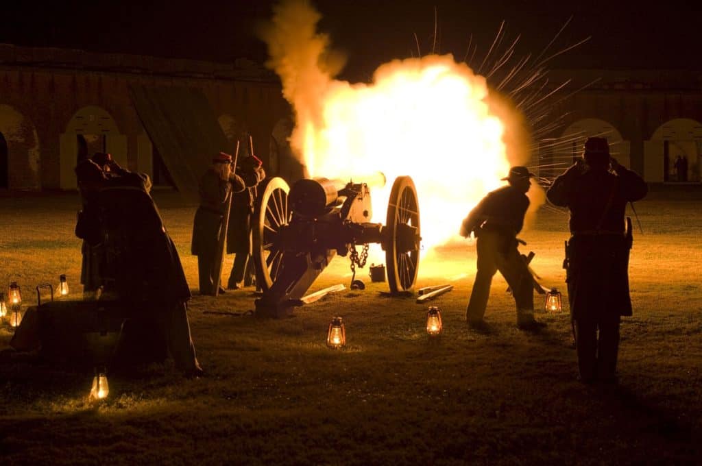 Five Civil War reenactors cover their ears at the blast of a canon. The canon fire lights up the night. 