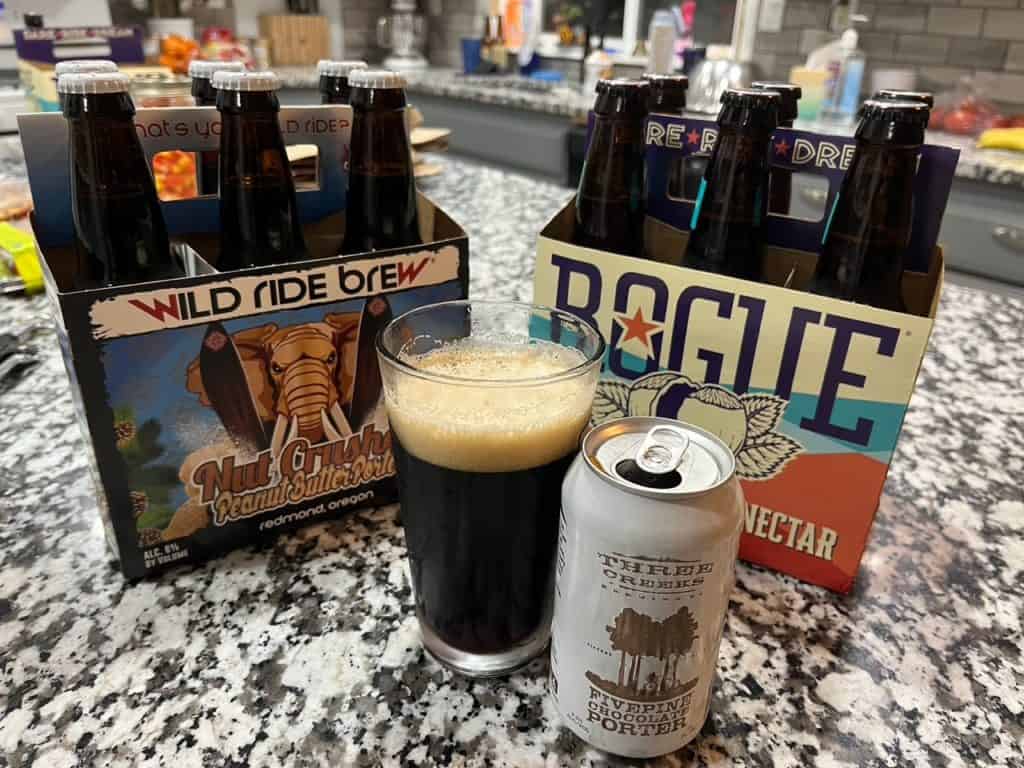 A tall glass of Five Pine Chocolate Porter stands in front of a six pack of Rogue Hazelnut Ale and a six packof WRB's Peanut Butter Porter. Craft beer is of one the best gifts fro Oregon.