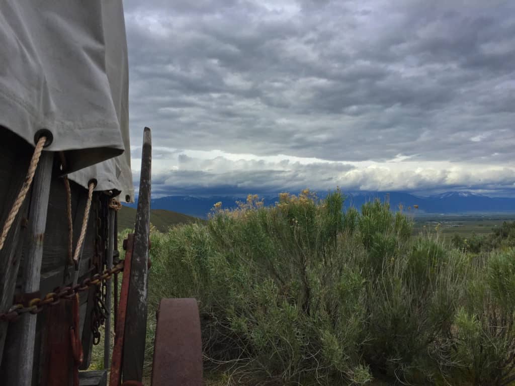 A covered wagon stands in a sage covered stretch of Eastern Oregon. Snowy, cloud-covered mountains stand on the horizon. 