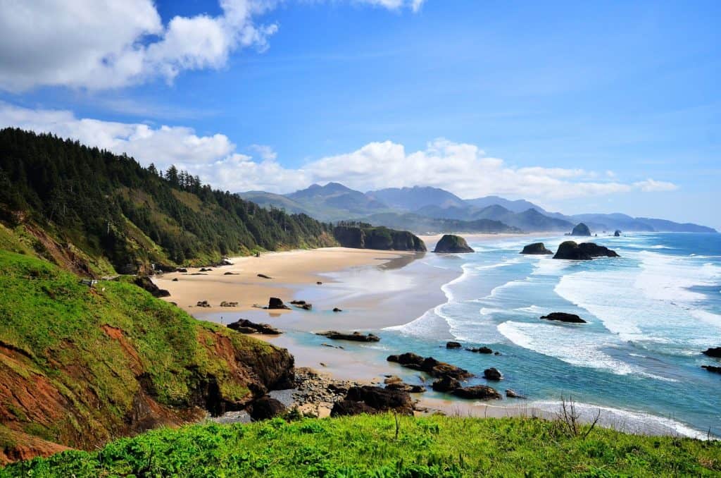 Blue ocean waves move about noble sea stacks and forested headlands at Cannon Beach and Ecola State Park. 