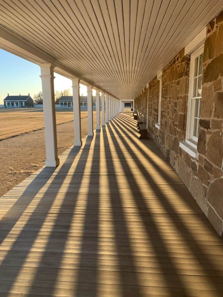 Morning sunlight casts long shadows along a columned porchway at Fort Larned National Historic Site. You can find several historic frontier sites on Dinkum Tribe's list of national parks and monuments by state. Image from NPS.