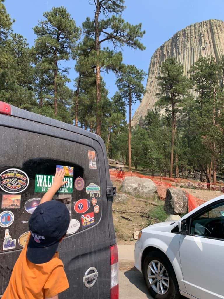 Our son places our latest bumper sticker on our van. The sticker says "DVL TWR". Devils Tower stands in the background. 
