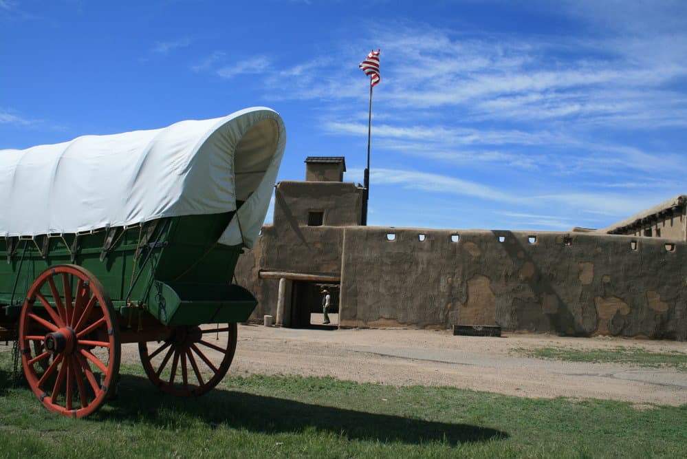 A Conestoga wagon stands in front of the adobe walls of Bent's Old Fort at Bent's Old Fort National Historic Site. You can discover historic sites and historic trails on Dinkum Tribe's List of National Parks and Monuments by State.