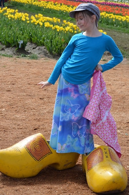 Girl standing in two giant wooden shoes in front of tulip field. Tulip Festival Oregon best time to go.
