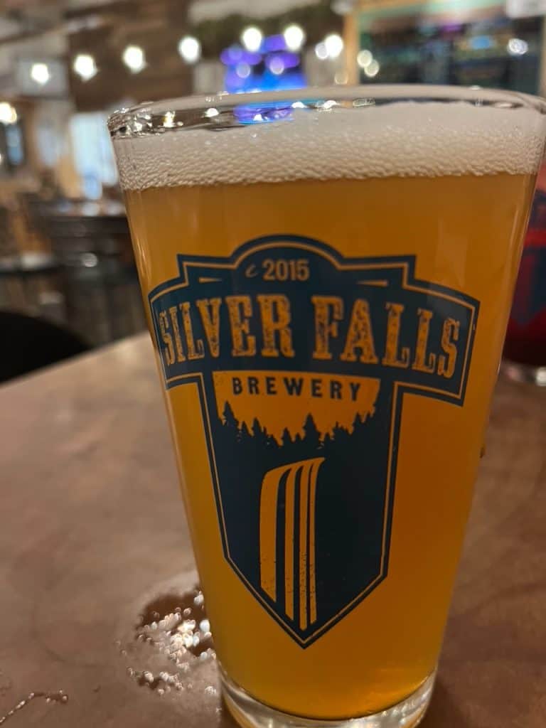 A tall glass of The Standing Hazy IPA stands on a table. The glass bears the logo of Silver Falls Brewery.