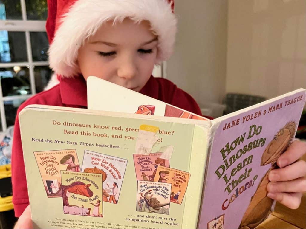 Boy in Santa hat reading How Do Dinosaurs learn Their Colors? book. Dinosaur gifts for a 5 year old.