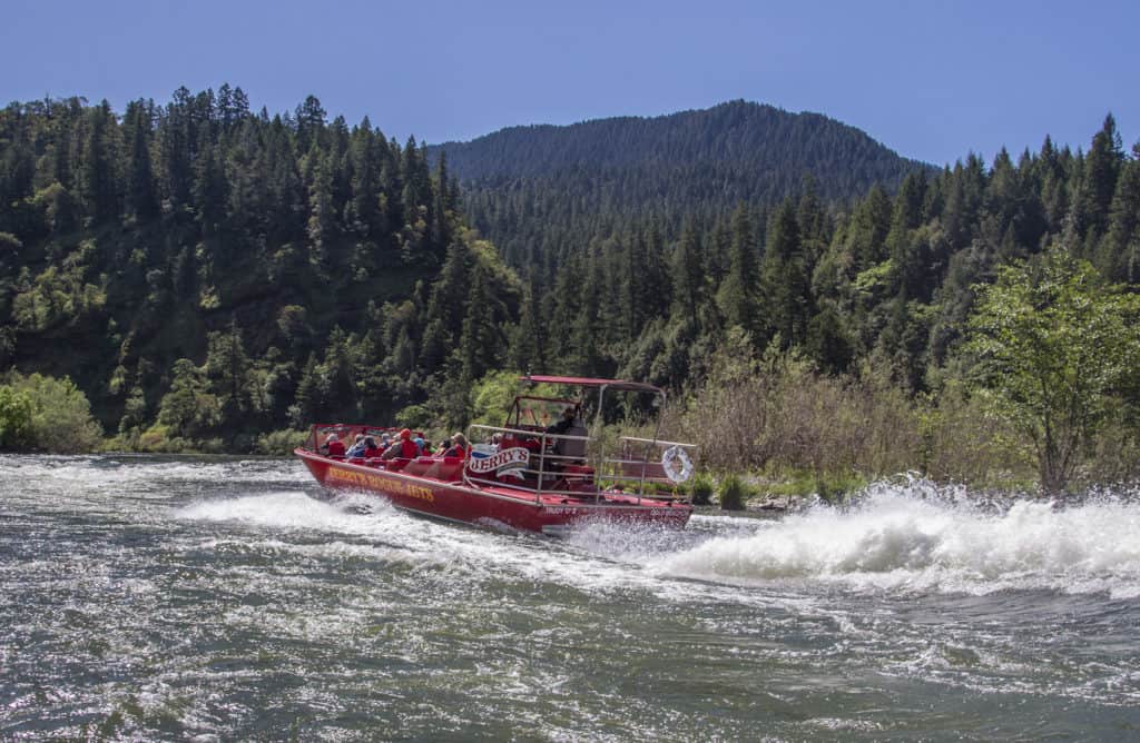 The image shows a red mail-boat full of visitors making its way up the Rogue River. 