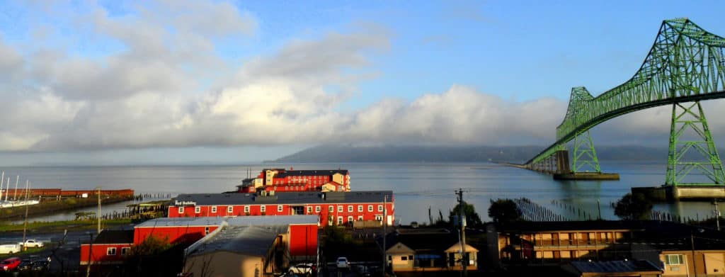The mouth of the Columbia River is framed by the green Astoria-Megler Bridge, red waterfront buildings, and the green slopes of Southern Washington.