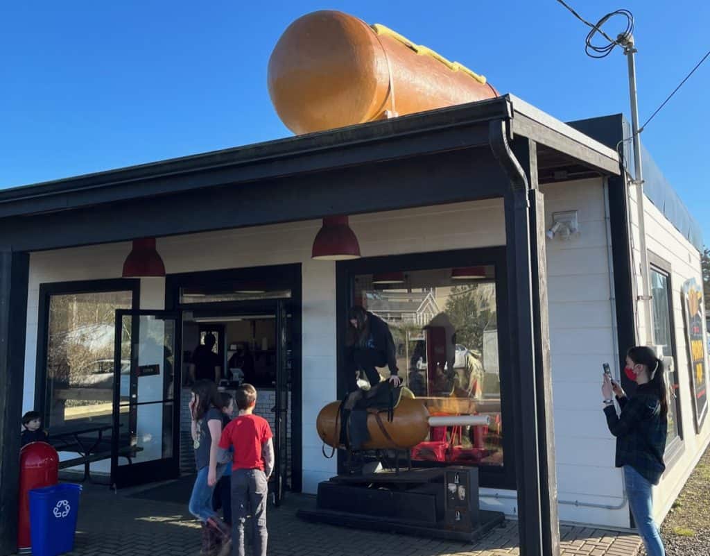 My teenage daughter prepares to ride the corn dog machine at Pronto Pup. We all stand near the entrance of the food stand that's topped by an enormous corn dog.