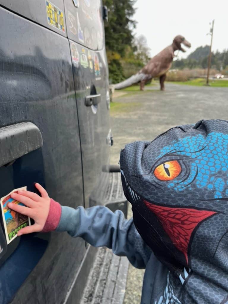 My son (dressed in a raptor sweater) sticks the Prehistoric Gardens bumper sticker on the back of our van. 