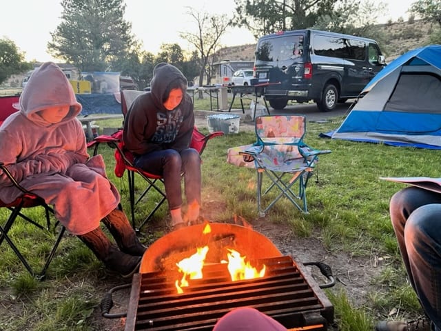 Girls by a fire in camping chairs with an empty kids chair next to them.