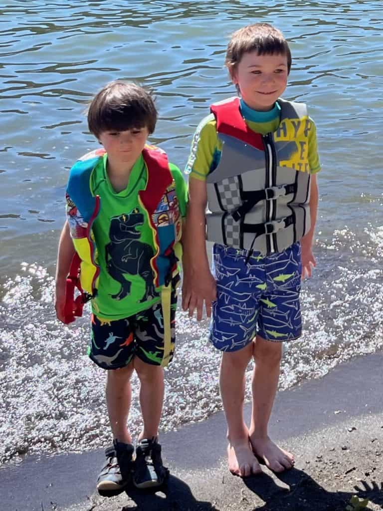 Two boys in front of lake wearing rash guards and life vests.