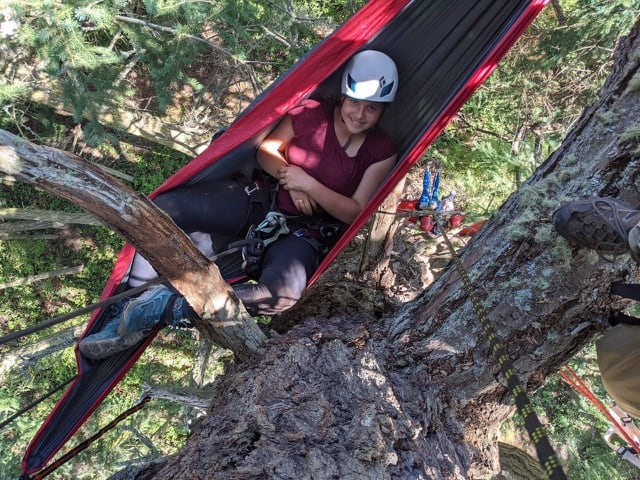 A young woman smiles from her hammock. The hammock is hanging way up at the top of a tree.