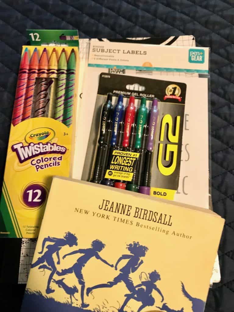 Homeschool supplies for a 6th grader: colored pencils, colored pens, writing notebook, fiction book, subject label stickers. 