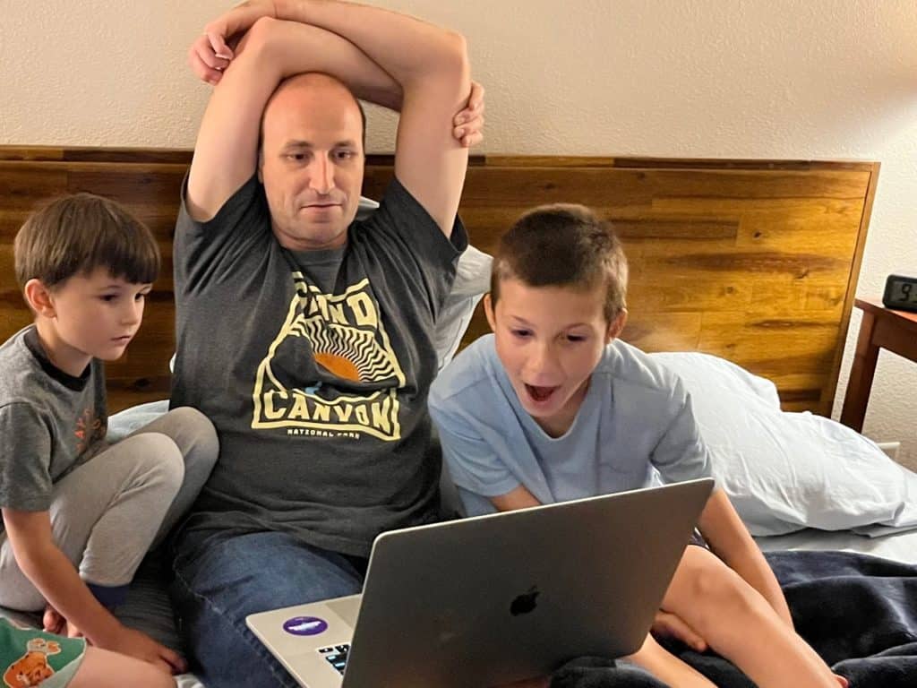Dad and boys watching computer on bed.