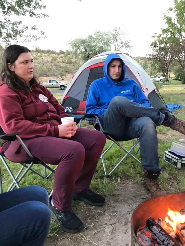 Man and woman by a campfire with cup in hand.