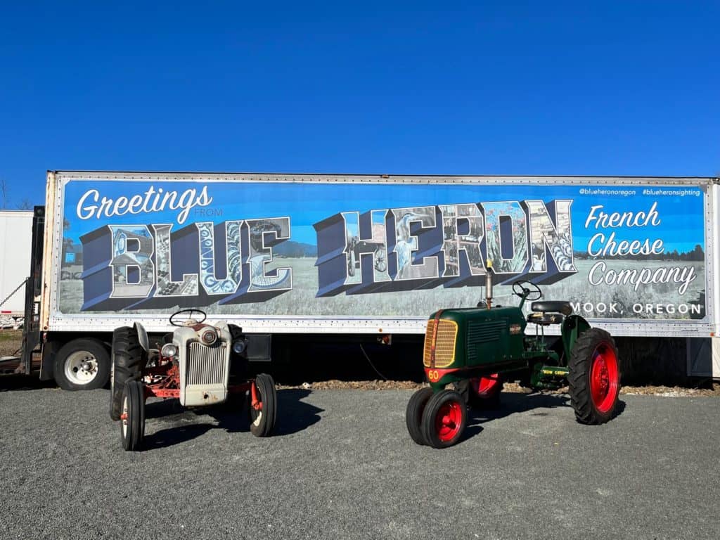 Tractors in front of a post card mural that says "Greetings from Blue Heron French Cheese Company" Tillamook, Oregon.