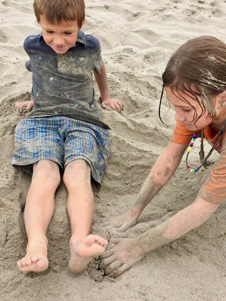 Girl burying a boy in the sand at the Seaside Beach.