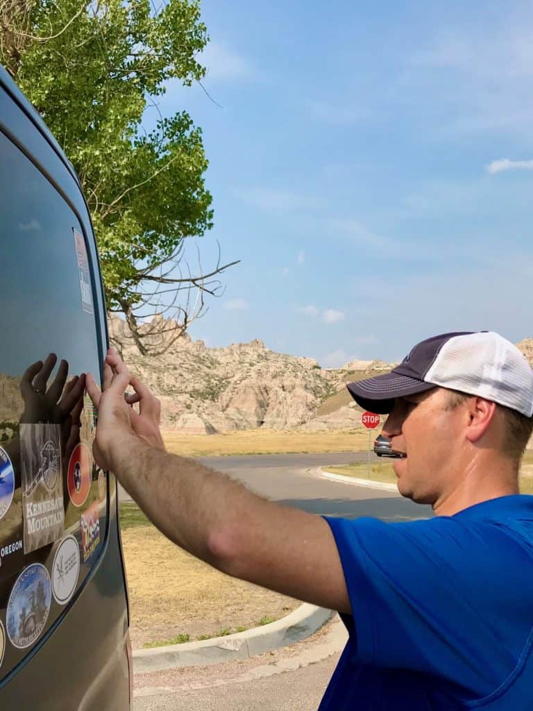 Man putting bumper sticker on van rear window with Badlands hills in the background.