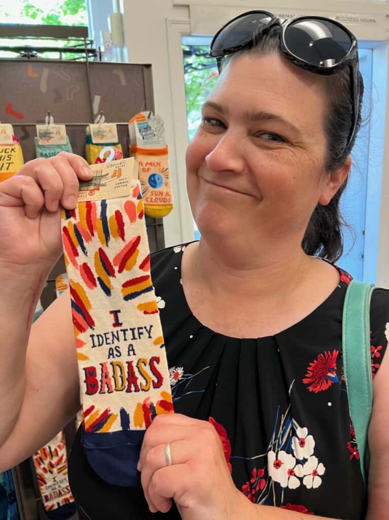 Jenn holding a pair of silly socks that say "I identify as a Badass"