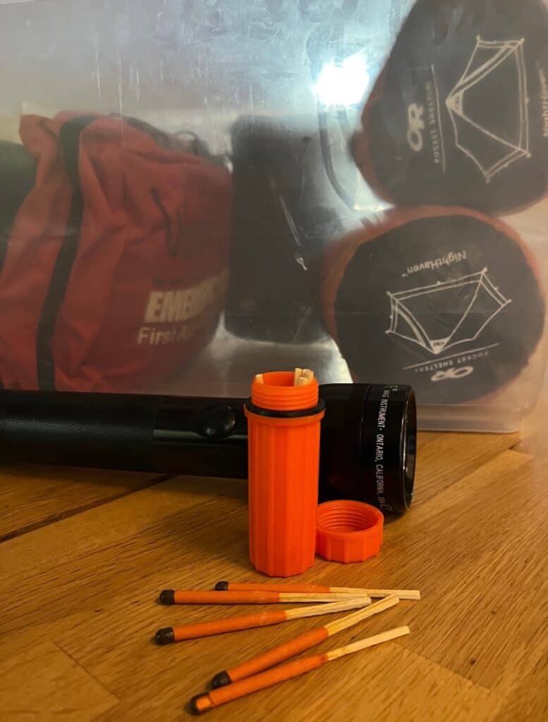 Waterproof matches and a Mag Light sit in front of a container filled with emergency supplies. Every motorist should take time to learn what to pack in a car emergency kit.