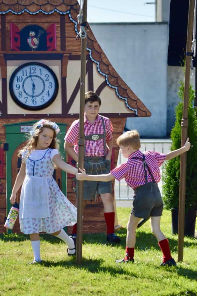Boys and girl in traditional Bavarian clothing dancing around flag poles.