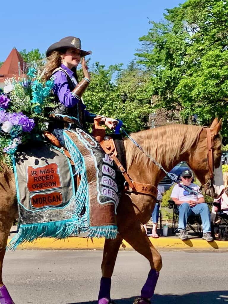 Girl on horseback in a parade. Her saddle blanket and chaps say, "Jr Miss Rodeo Oregon".