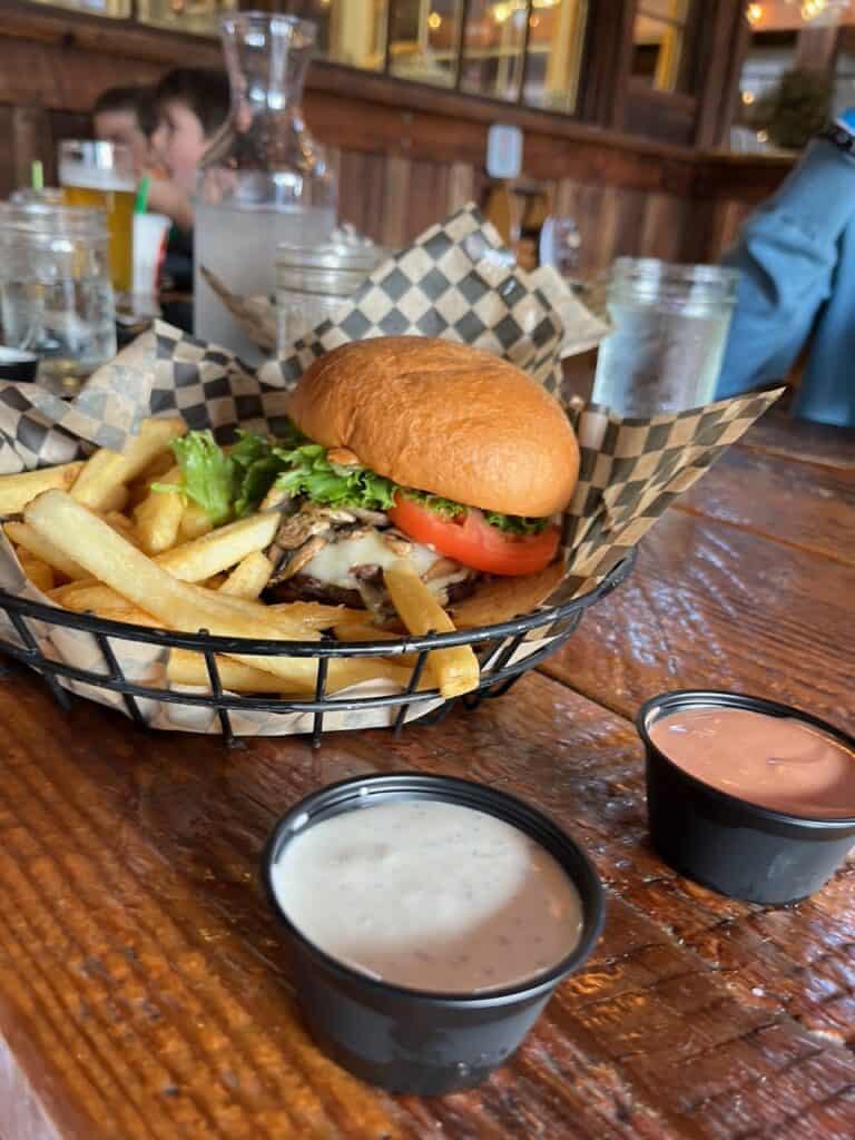 A Mushroom Swiss burger with a side of fries sits beside dipping sauces. Grain Station Brew Works is one of the best breweries in McMinnville and Carlton.