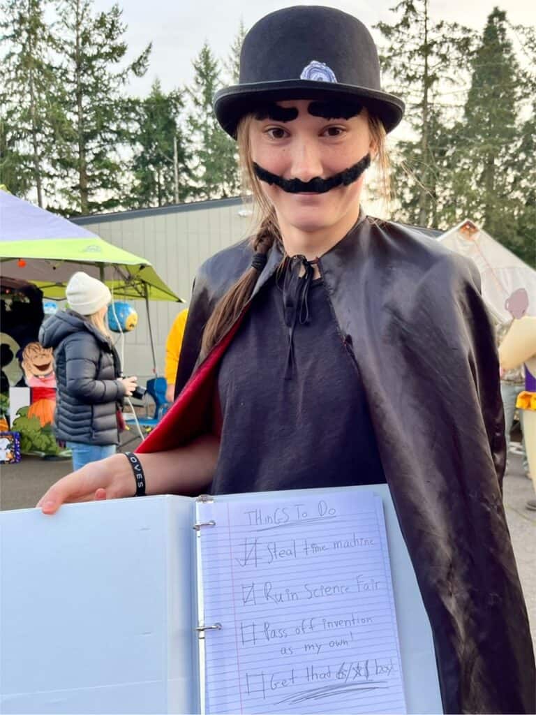 Our teen daughter in a bowler hat, fake mustache, and black cape with a binder and checklist. Her costume is the Bowler Hat Guy from the movie Meet the Robinsons.