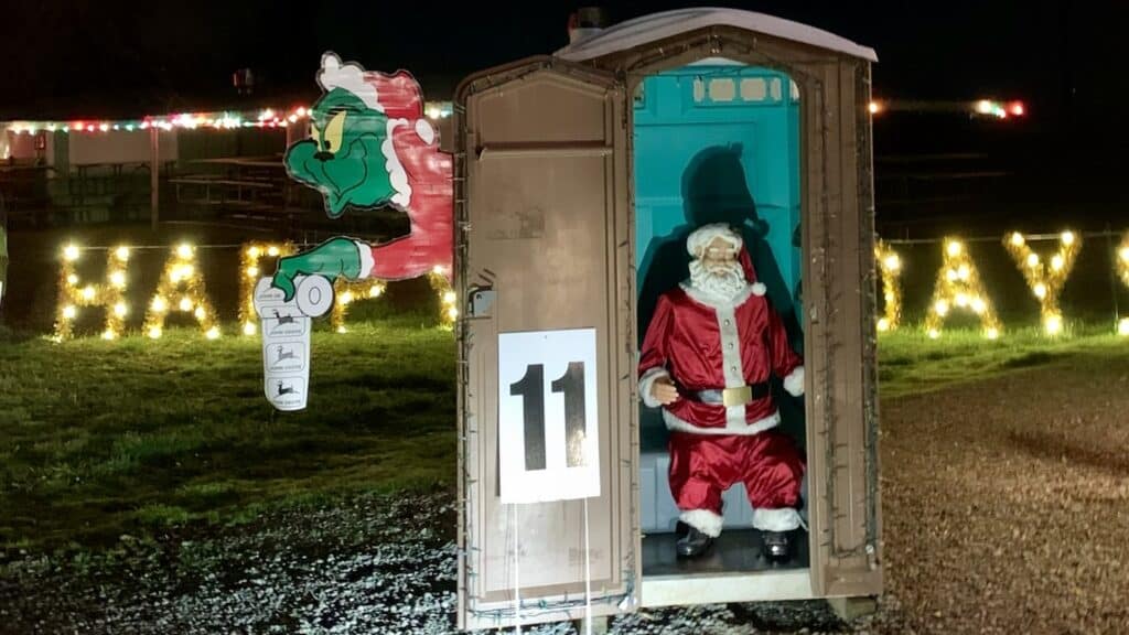 Santa mannequin in a portable toilet with the Grinch outside holding John Deere monogrammed toilet paper. 