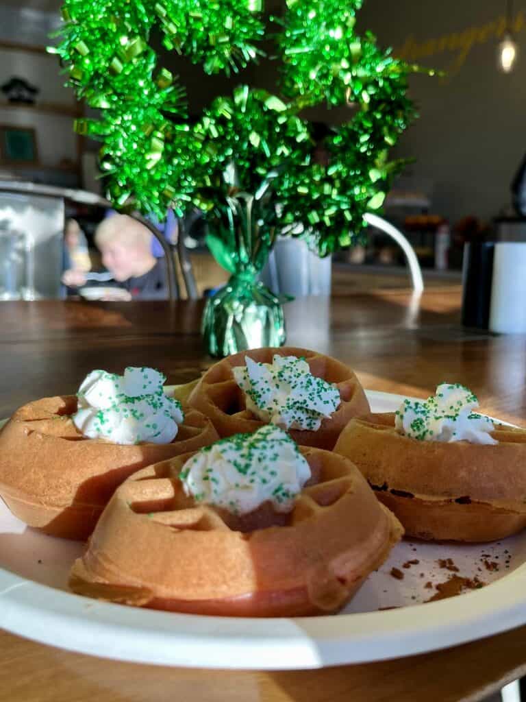 Four pink mini waffles with whipped cream and green sugar sprinkles with a green shamrock in the background.