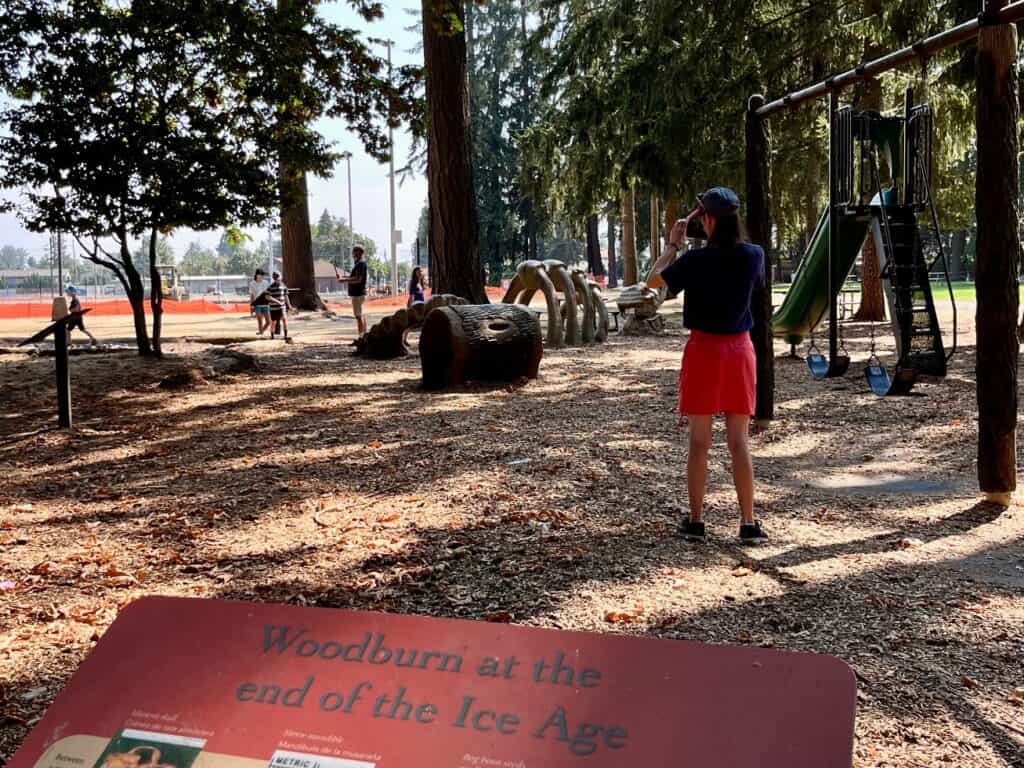 Our daughter looks towards the play area at Legion park. An interpretive sign in the foreground is titled "Woodburn at the End of the Ice Age".