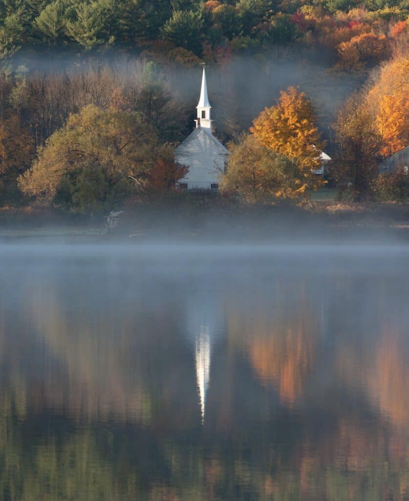 A misty lake reflects a white church steeple on an autumn morning in New Hampshire.