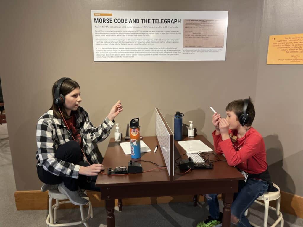 Teen girl and boy at a table with headphones sending messages by telegraph using Morse Code