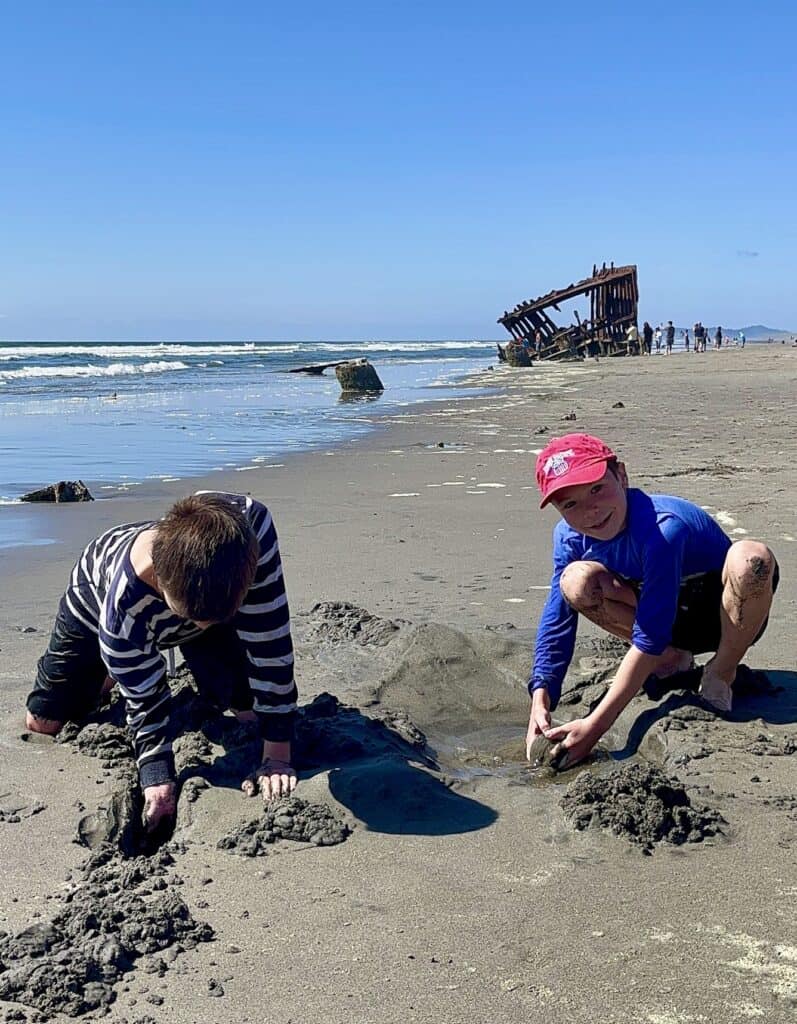 A shipwreck stands in the background as out boys build sandcastles at Lewis & Clark National Historical Trail, Oregon.